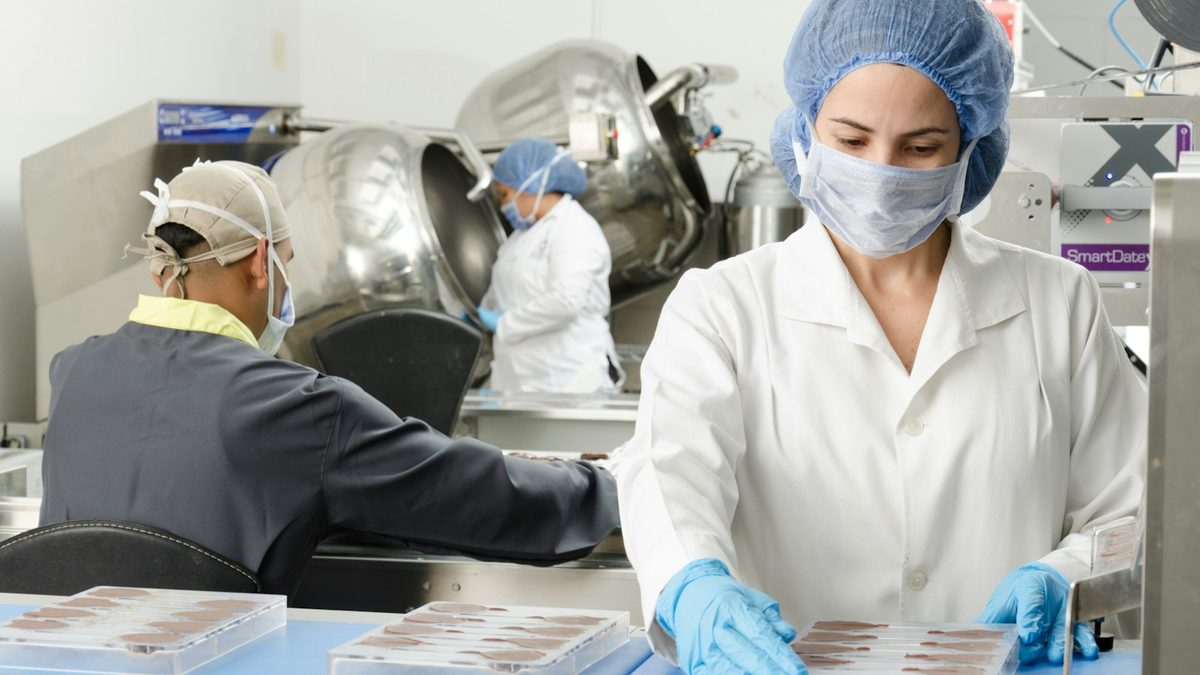 Workers in hairnets and masks packaging product trays on a production line next to a date-coding printer