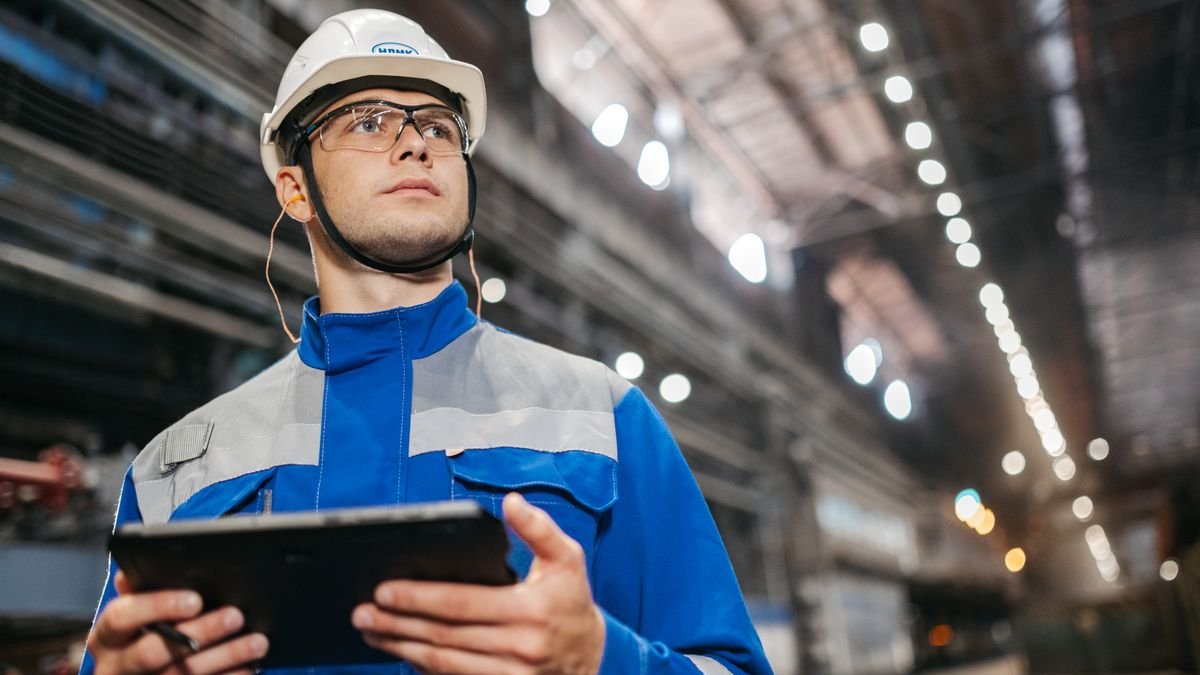 Industrial worker reviewing equipment data on a tablet inside a large manufacturing facility